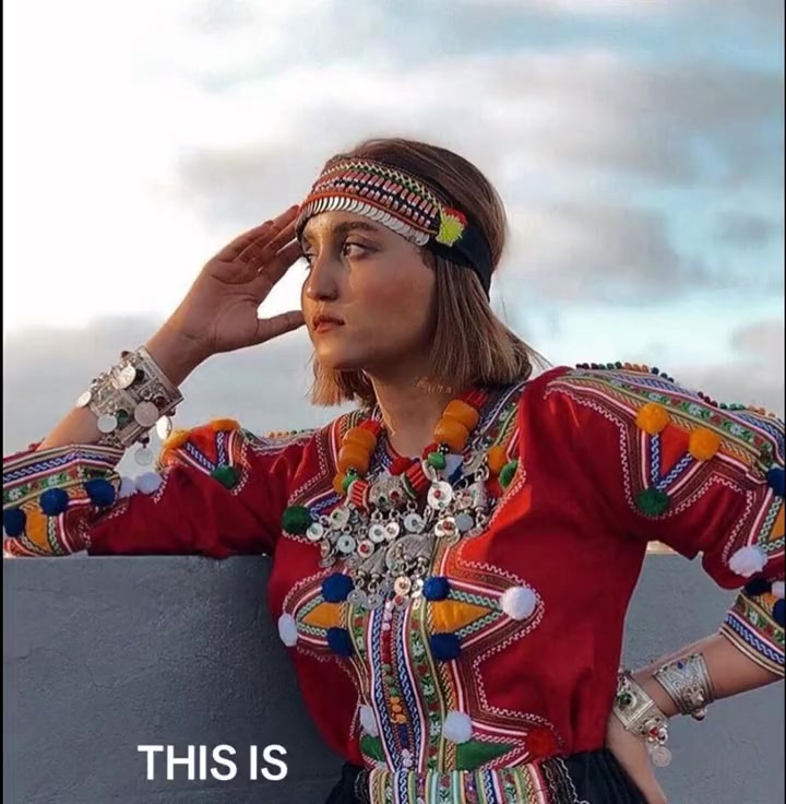 A young Algerian woman in traditional Amazigh (Berber) indigenous dress — a vivid red embroidered top with colorful pompoms, silver coin jewelry, and a beaded headband. Face uncovered.