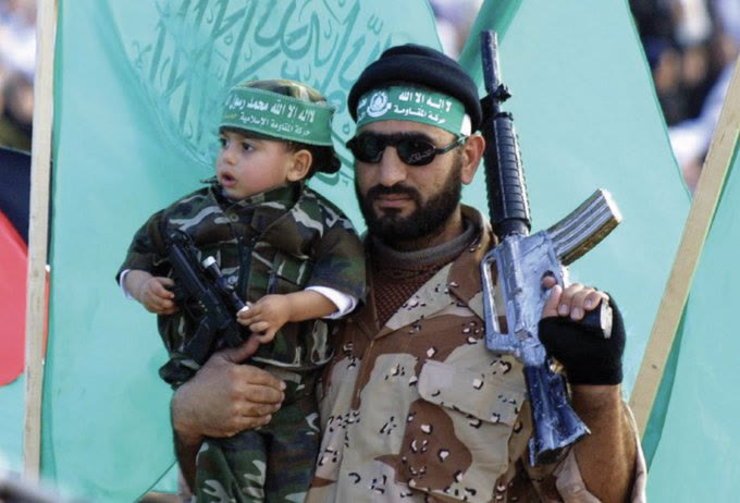 A Hamas fighter in military fatigues and green Hamas headband holds a toddler also dressed in camouflage with a matching Hamas headband. The fighter holds a rifle. Green Hamas flags are visible in the background at what appears to be a public rally.