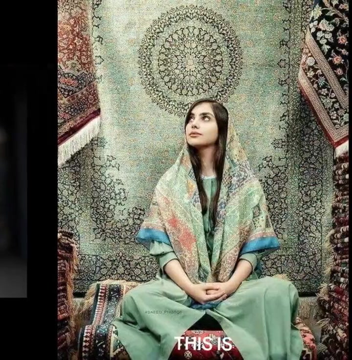 A young Iranian woman in traditional Persian dress — sage green garments with an ornate floral shawl draped loosely over her head, hair visible, surrounded by Persian carpets.